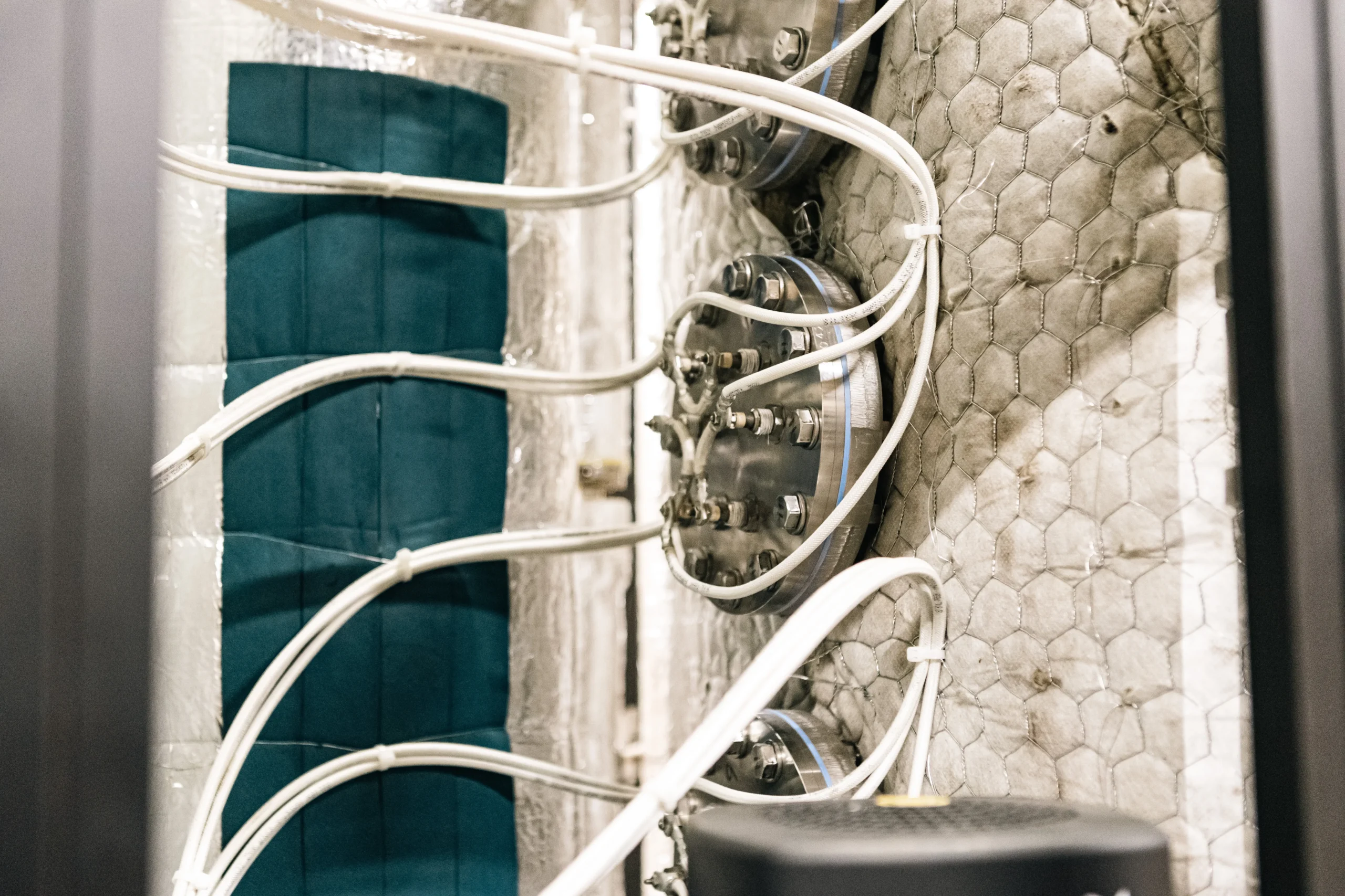 Ångledningar - PJP Piping Närbild av en industrimaskin med flera vita ångledningar kopplade till metalliska cirkelformade komponenter, mot en texturerad, hexagonmönstrad yta.